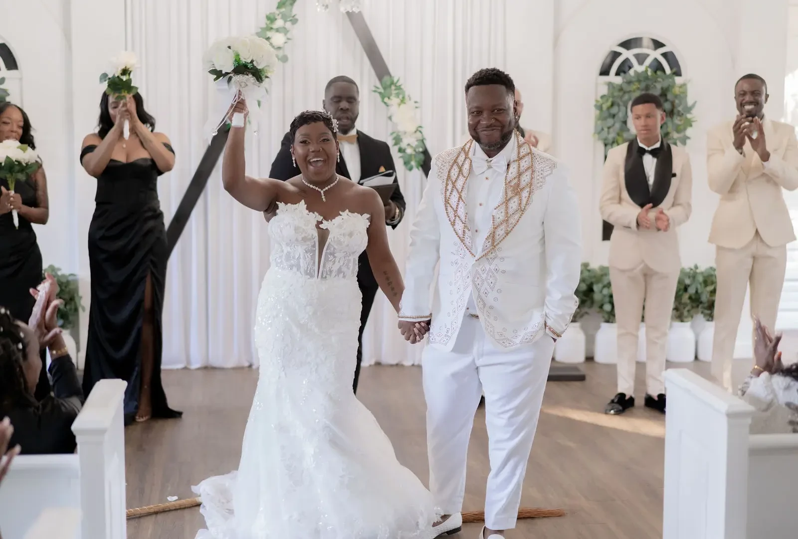 A newlywed couple smiles and holds hands while walking down the aisle after their wedding ceremony, surrounded by guests celebrating.