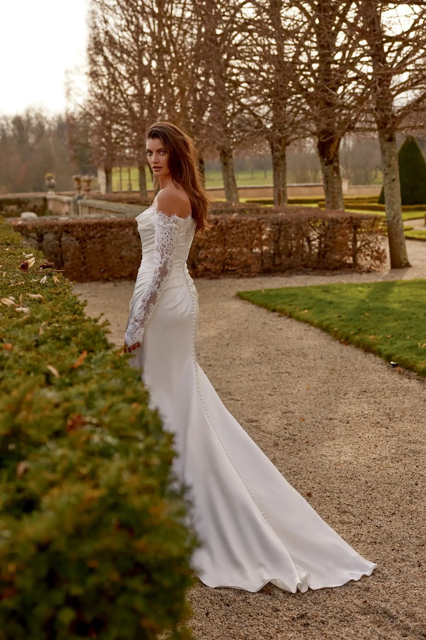 Bride in an elegant, off-shoulder lace wedding dress standing on a gravel path, surrounded by bare trees and manicured hedges in a serene garden.
