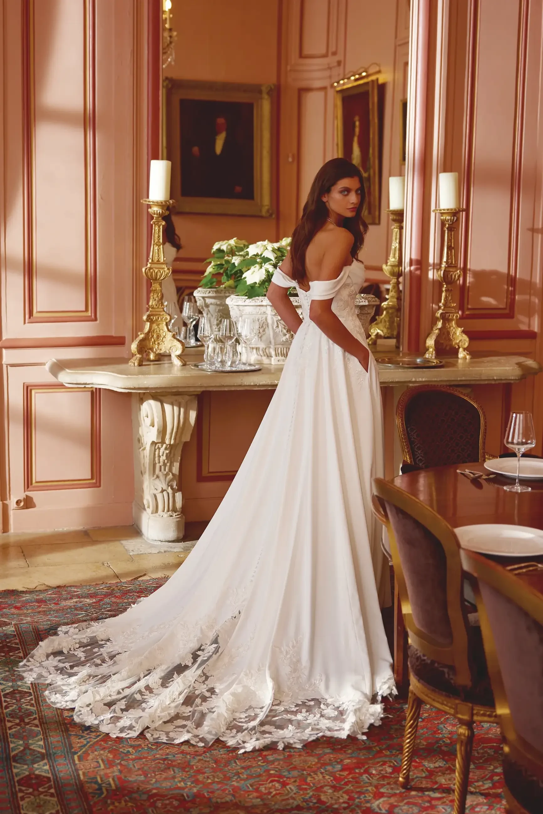 A woman in an elegant off-the-shoulder white wedding gown stands in a luxurious room. The dress, which has a long, lace-edged train, adds a touch of sophistication. She gazes back with a serene expression. The ornate room features a plush rug, candleholders, and classic portraits, creating a refined, regal atmosphere.