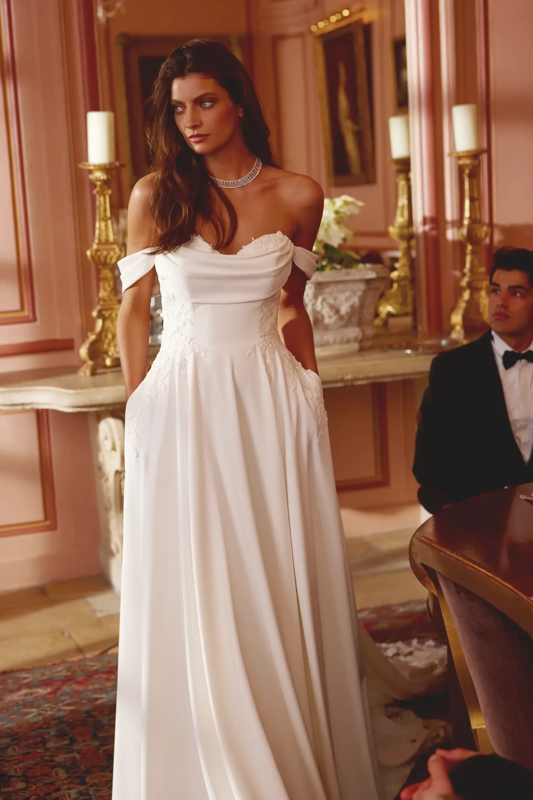 Bride in elegant, off-shoulder white gown with lace details stands in a luxurious, candlelit room. She looks contemplative; a man in a tuxedo sits nearby.