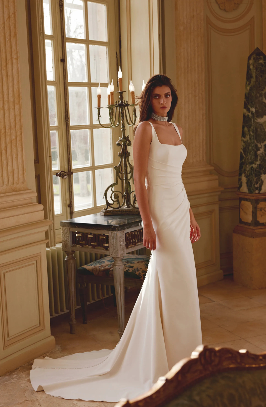 A woman in a sleek, white gown with a long train stands in an elegant room with large windows and candleholders, exuding an aura of sophistication.