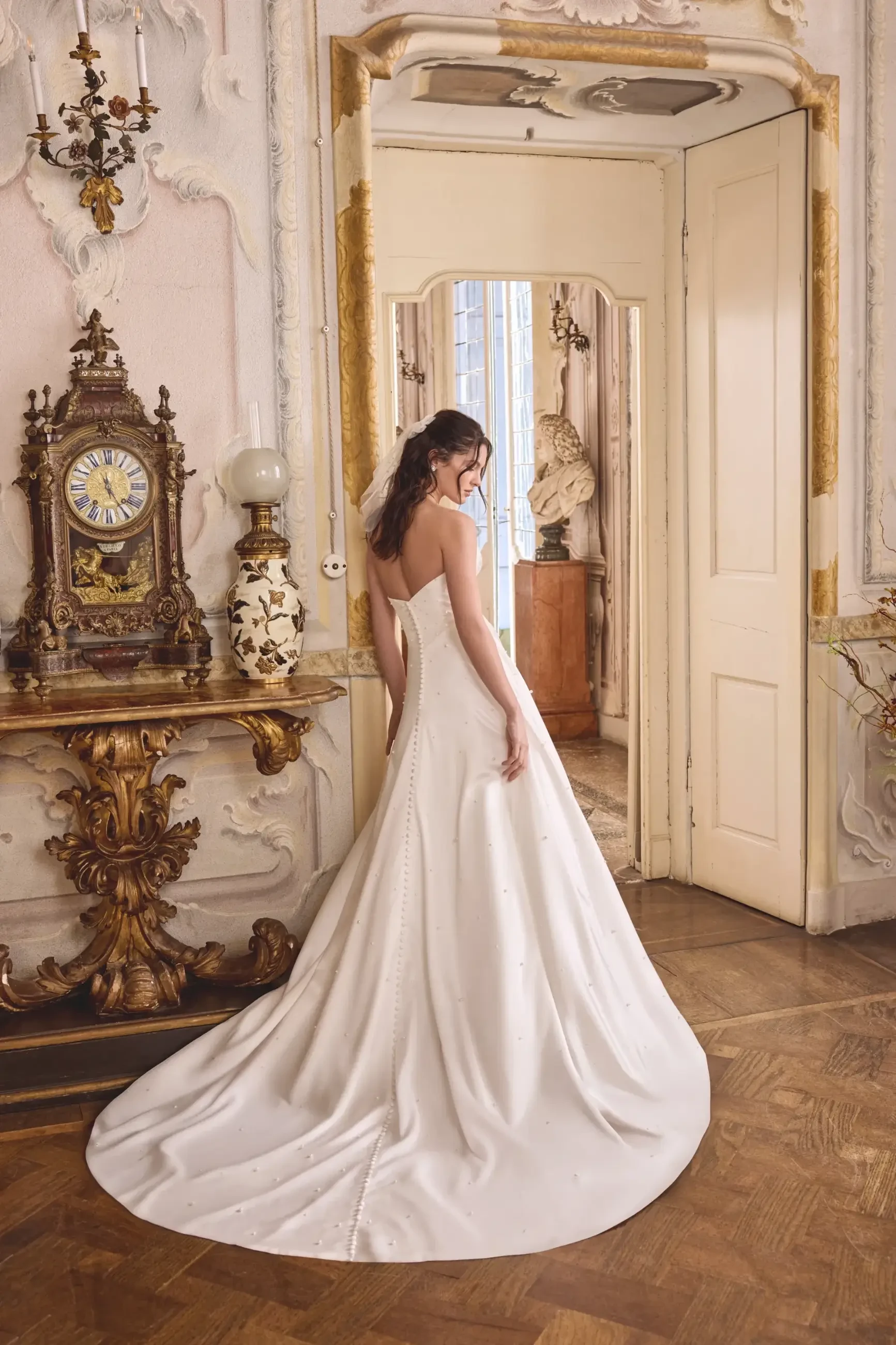 A bride in an elegant white gown stands in an ornate, sunlit room. The dress has a fitted bodice and flowing skirt, exuding grace and sophistication.