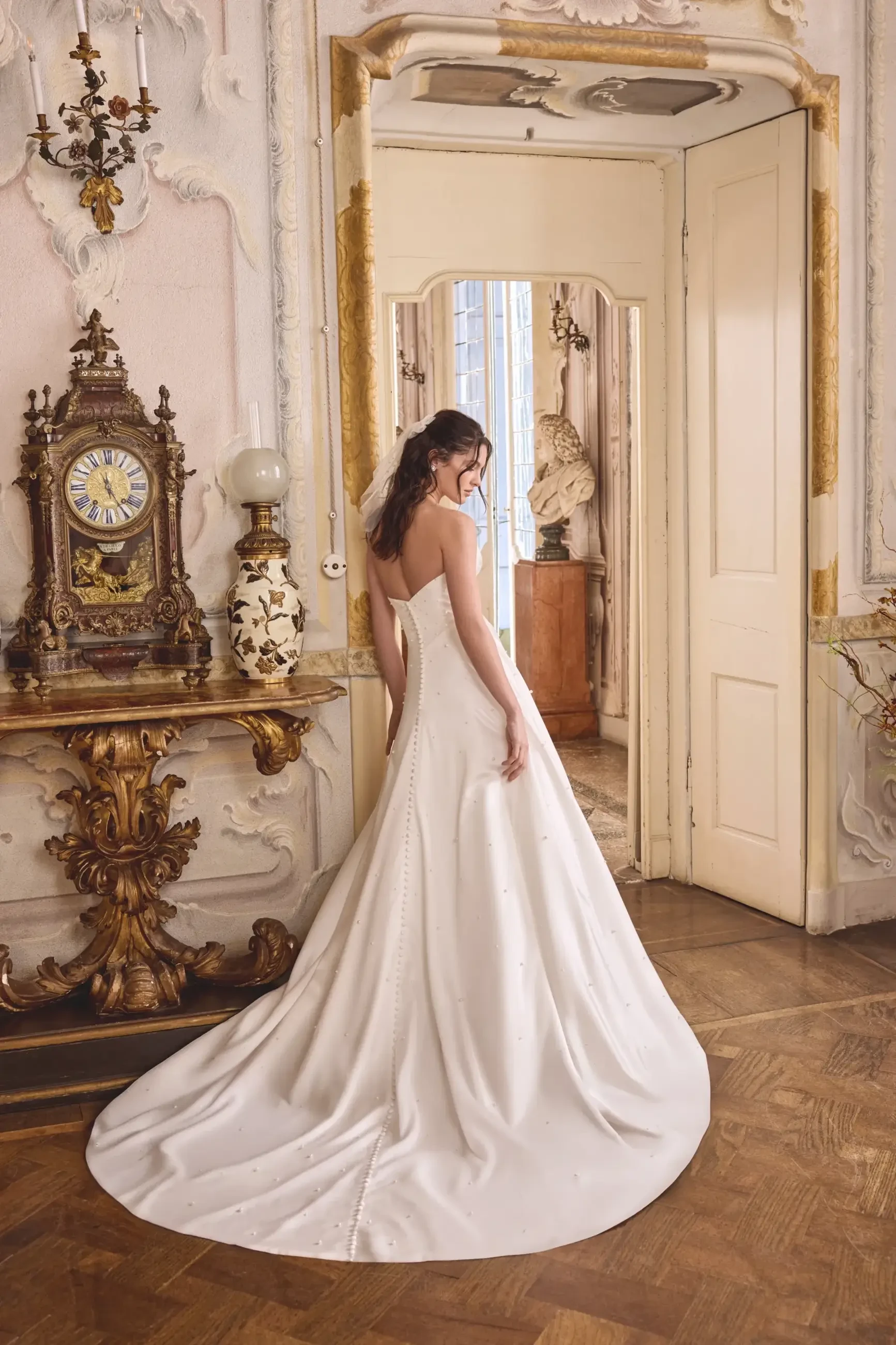 A woman wearing a white wedding dress stands with her back to the camera in an ornate room featuring elegant decor and wooden flooring.