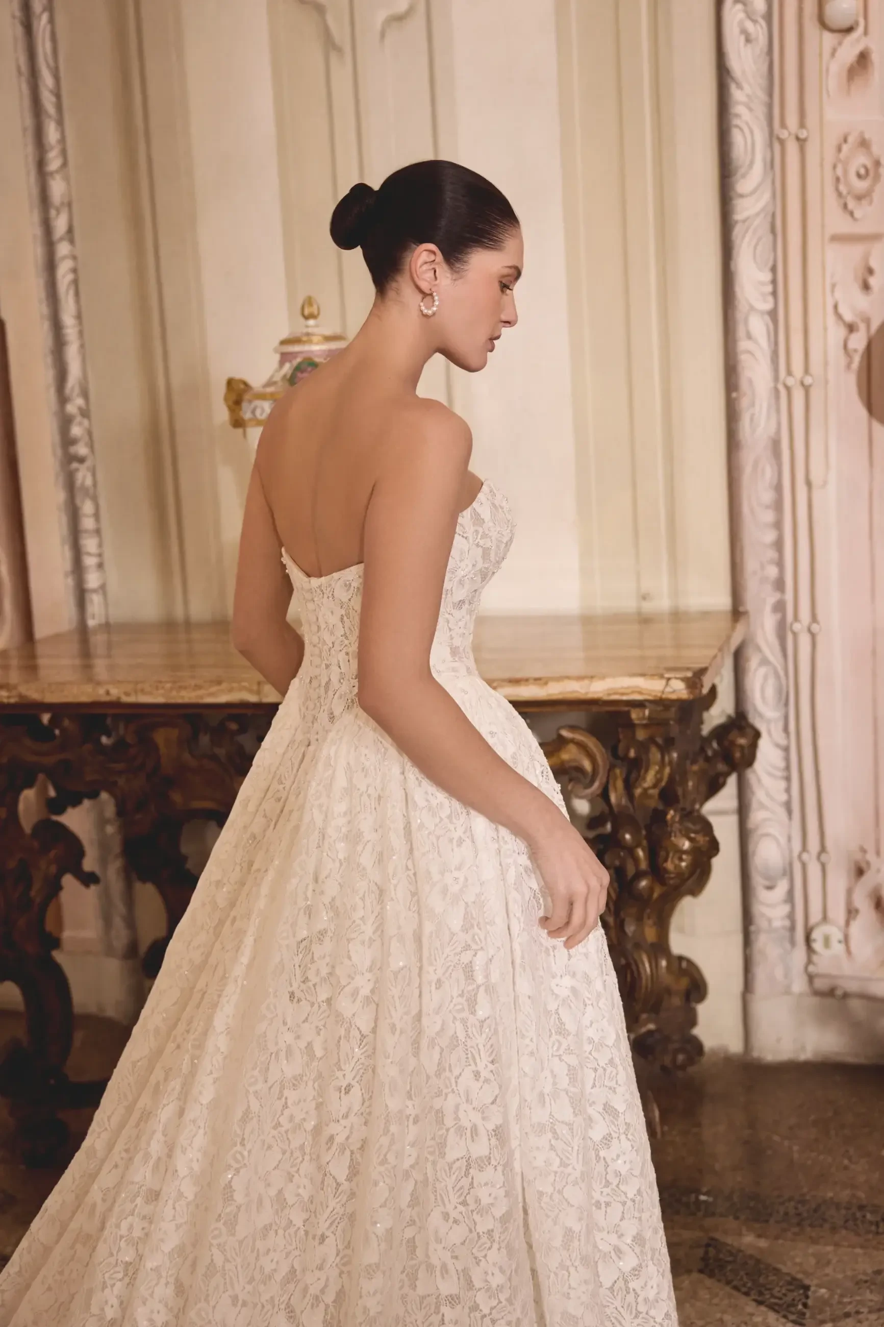 Elegant woman in a lace bridal gown stands in a luxurious room with ornate furniture, exuding sophistication and grace. The mood is serene and poised.