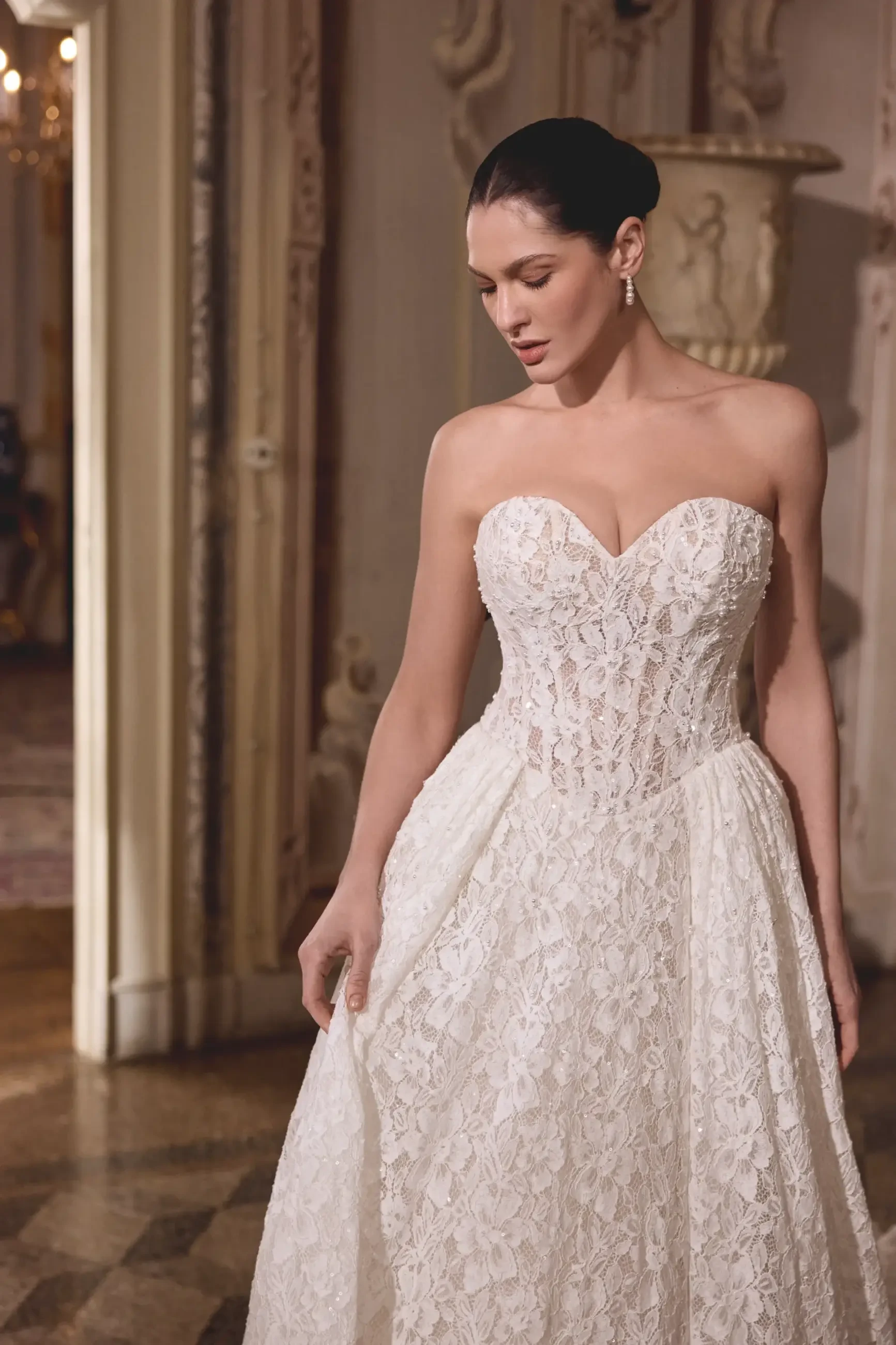 A woman in an elegant strapless lace wedding gown stands in a richly decorated room. Her calm expression and classic updo enhance the formal, romantic ambiance.