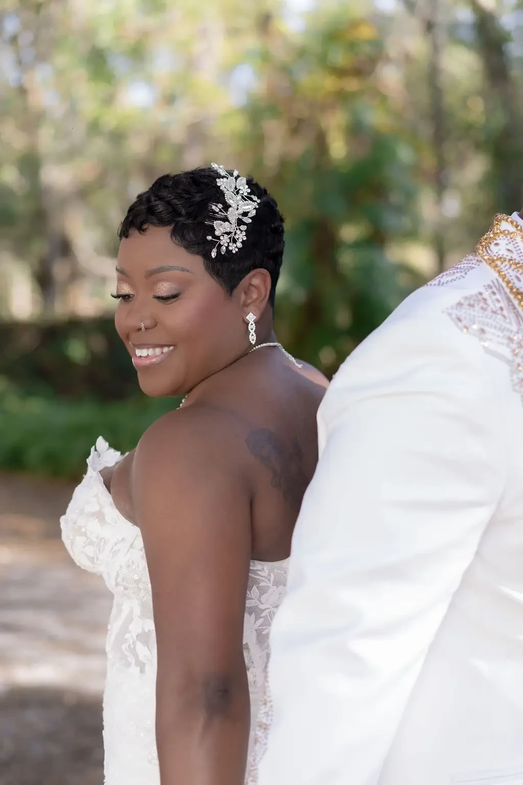 A smiling bride in a white wedding dress and hair accessory poses with her back to the camera, while the groom stands behind her, partially out of view.