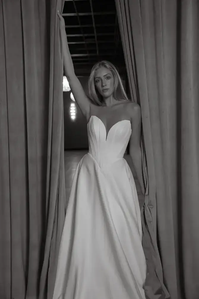 A woman in a white strapless gown stands between two curtains, looking into the camera with a serious expression. The image is in black and white.