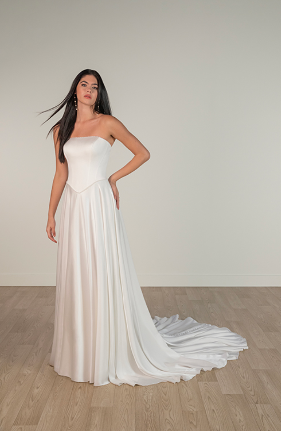 A woman in a strapless white wedding gown with a flowing train stands on a wooden floor. Her long, dark hair cascades down, conveying elegance and confidence.