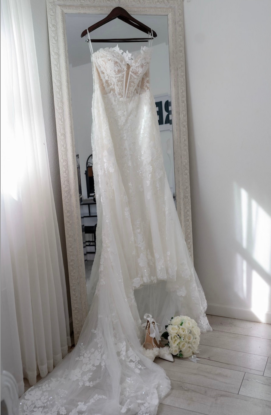 A wedding dress hanging in front of a mirror, with a pair of shoes and a bouquet at the base.
