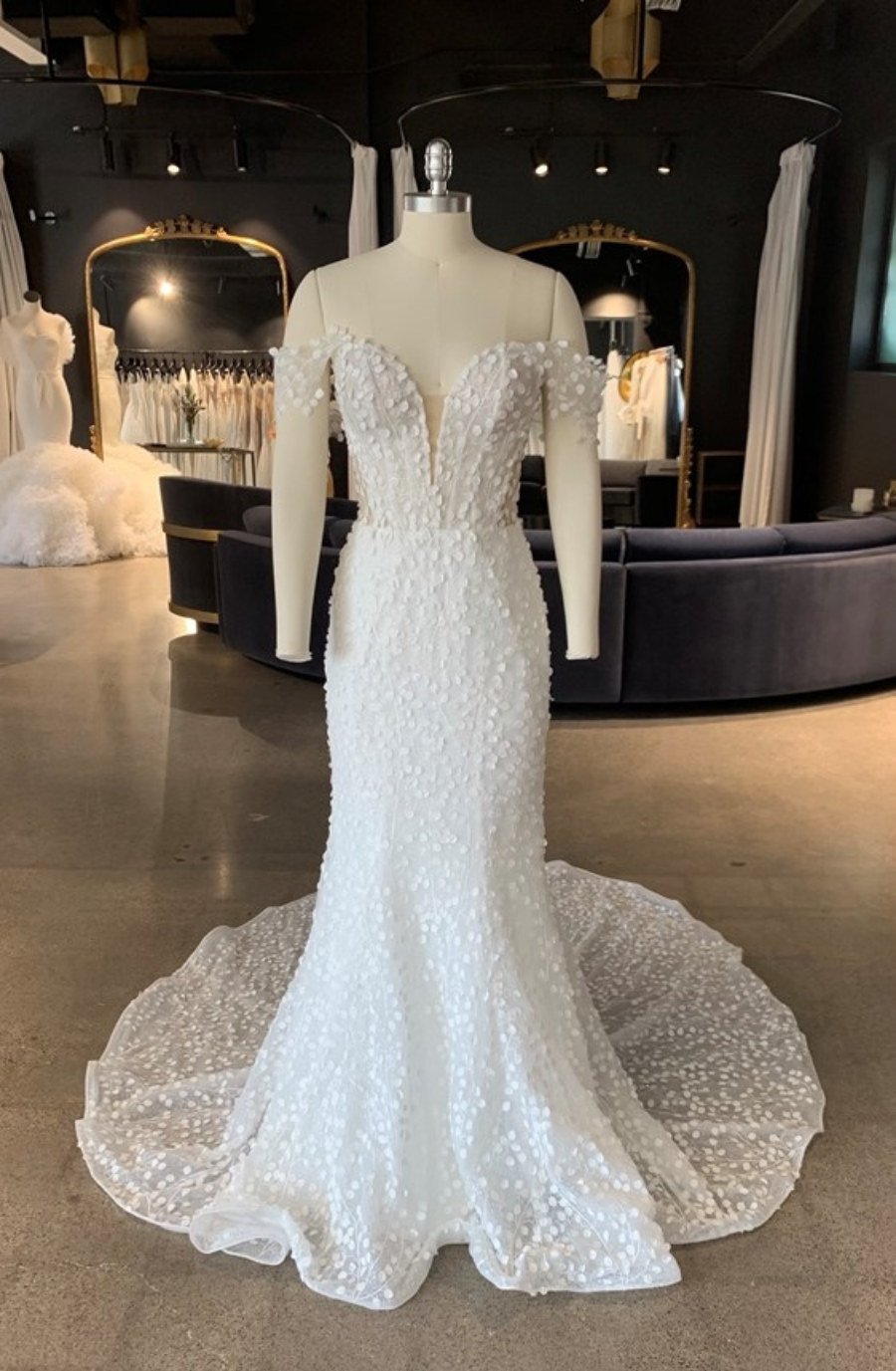 Elegant white wedding dress on a mannequin, featuring off-the-shoulder sleeves and floral appliqu&eacute;, with a flowing train in a chic boutique setting.