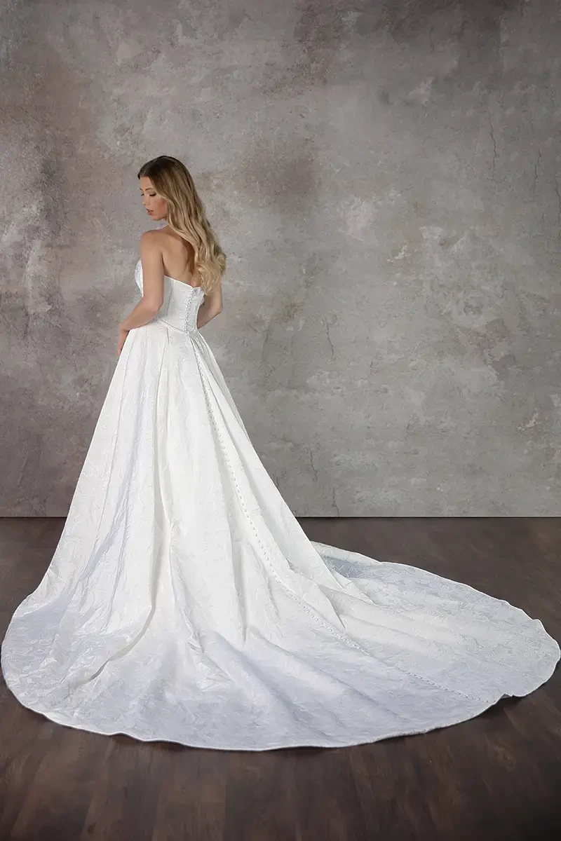 A woman in a strapless white wedding gown with a long train stands gracefully on a wooden floor against a textured gray backdrop, exuding elegance.