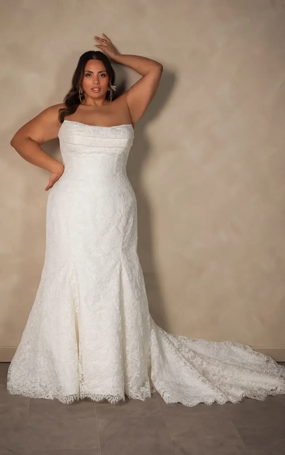 A plus-size woman in a strapless white wedding dress, standing against a neutral background with her hand on her head.