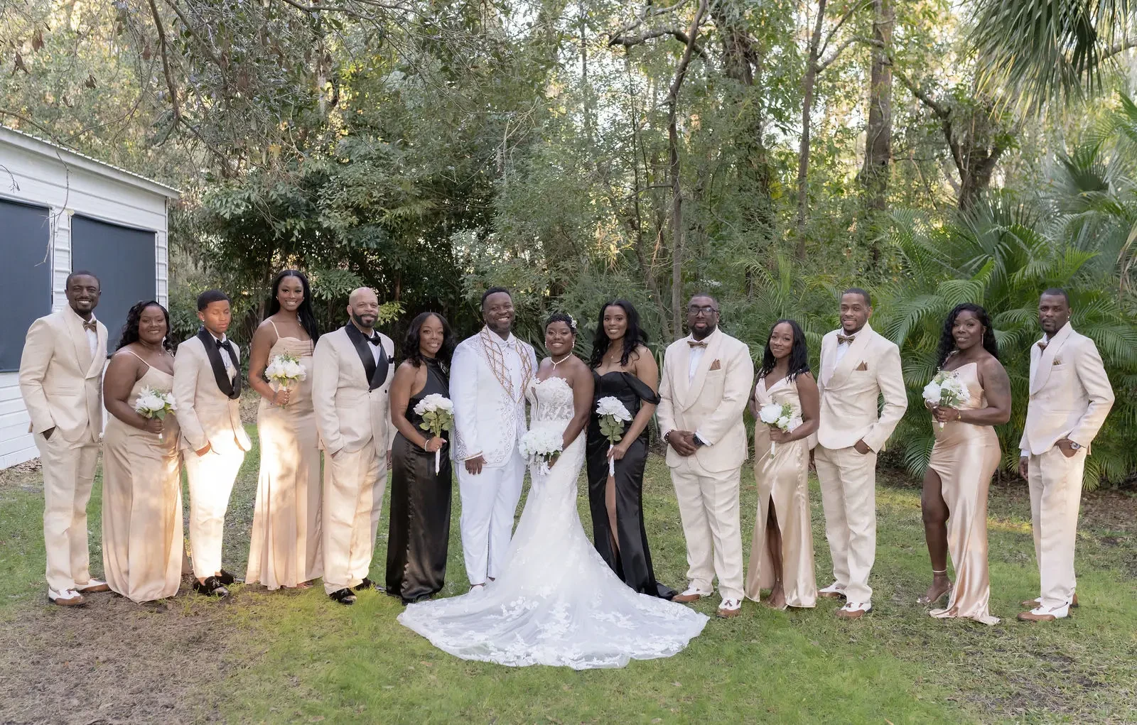 A wedding party poses for a group photo outdoors, surrounded by trees and greenery. The bride is wearing a white gown, while the groom is in a white suit. The wedding party members are dressed in various formal attire, including light-colored suits and elegant dresses.