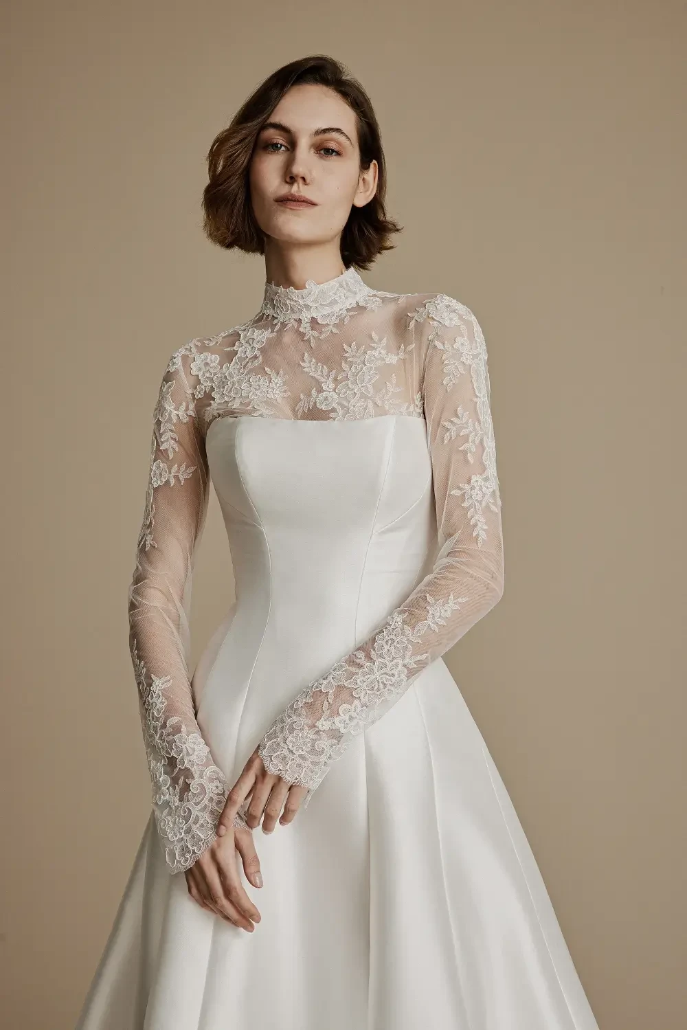 A woman in a fitted white wedding dress with long lace sleeves, standing against a beige background.