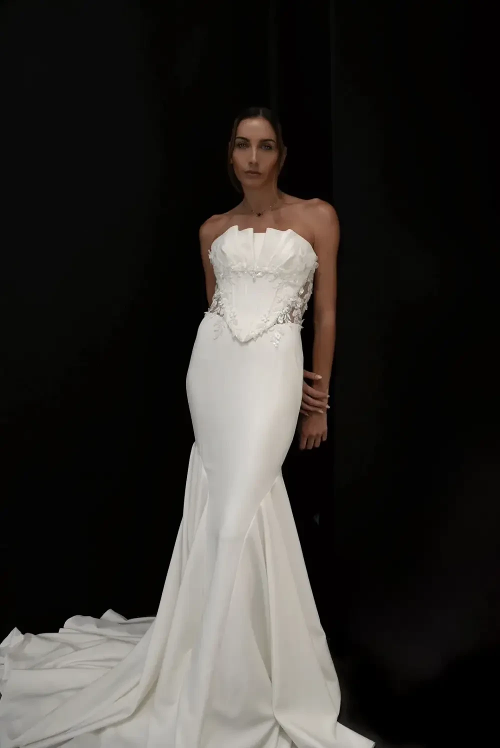 A model wearing a sleek, strapless white gown with intricate detailing stands against a black background.