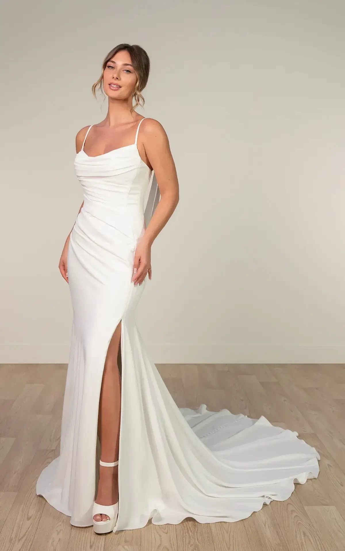 A woman is wearing a white formal dress with thin straps and a side slit, standing on a wooden floor with a neutral background.