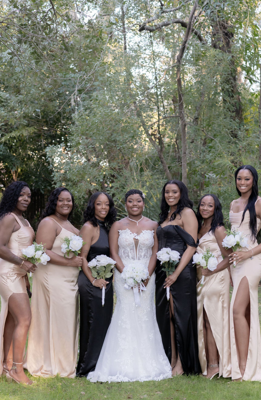 A bride stands in the center with seven bridesmaids, all wearing elegant dresses. The bride is in a white gown, and the bridesmaids wear various shades of gold and black. They are outdoors surrounded by greenery, smiling for the photo and holding bouquets of flowers.