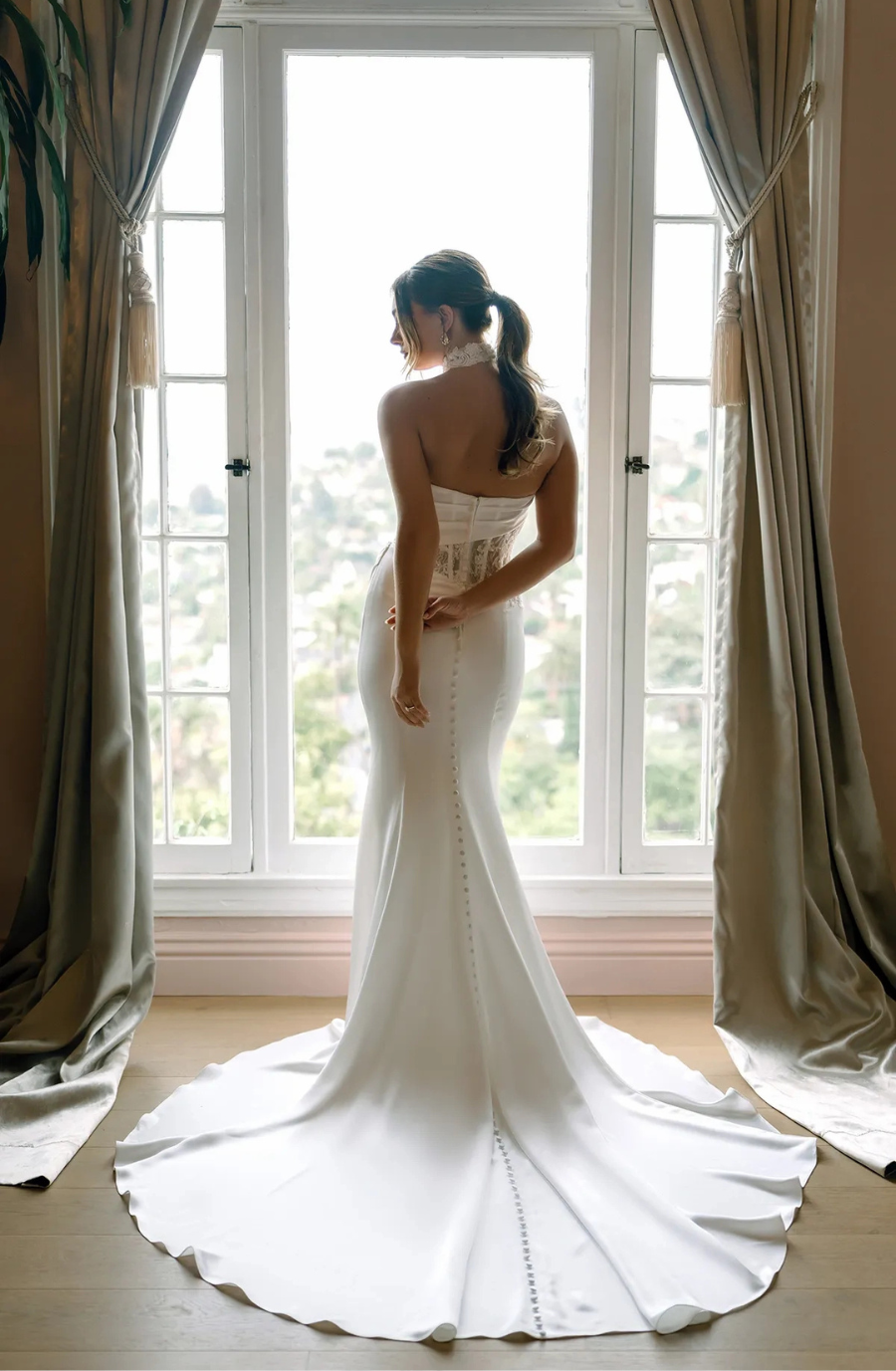 A woman in a white, fitted wedding dress with a long train stands by a large window. Sunlight filters through, creating a serene and elegant atmosphere.