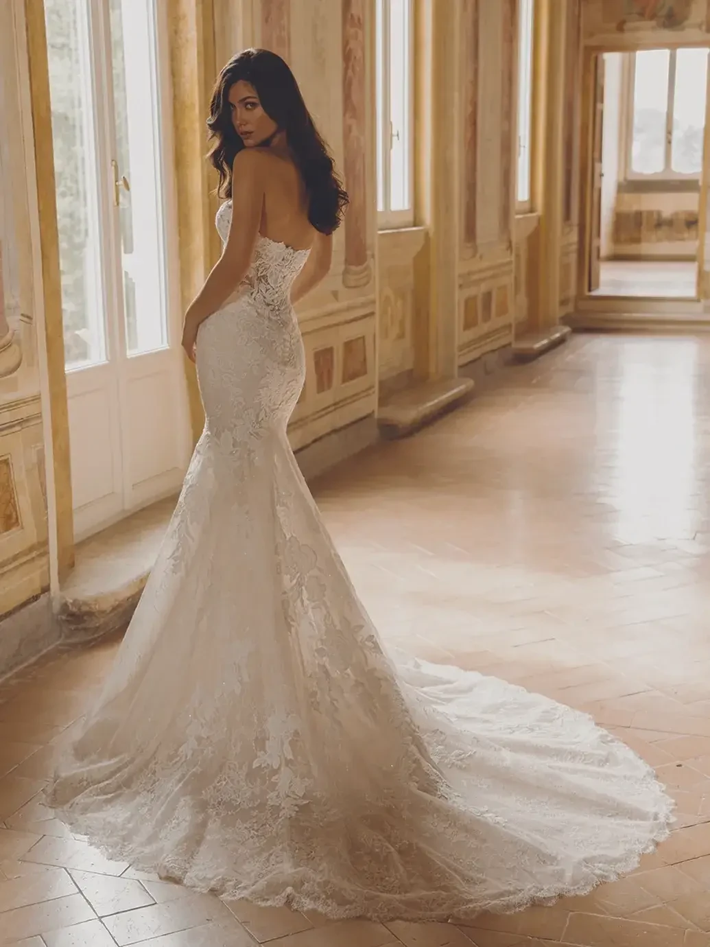 A woman wearing an elegant, strapless wedding gown with lace details stands in a beautiful, ornate room. The gown features a fitted silhouette and a long train, and the woman is looking back over her shoulder.