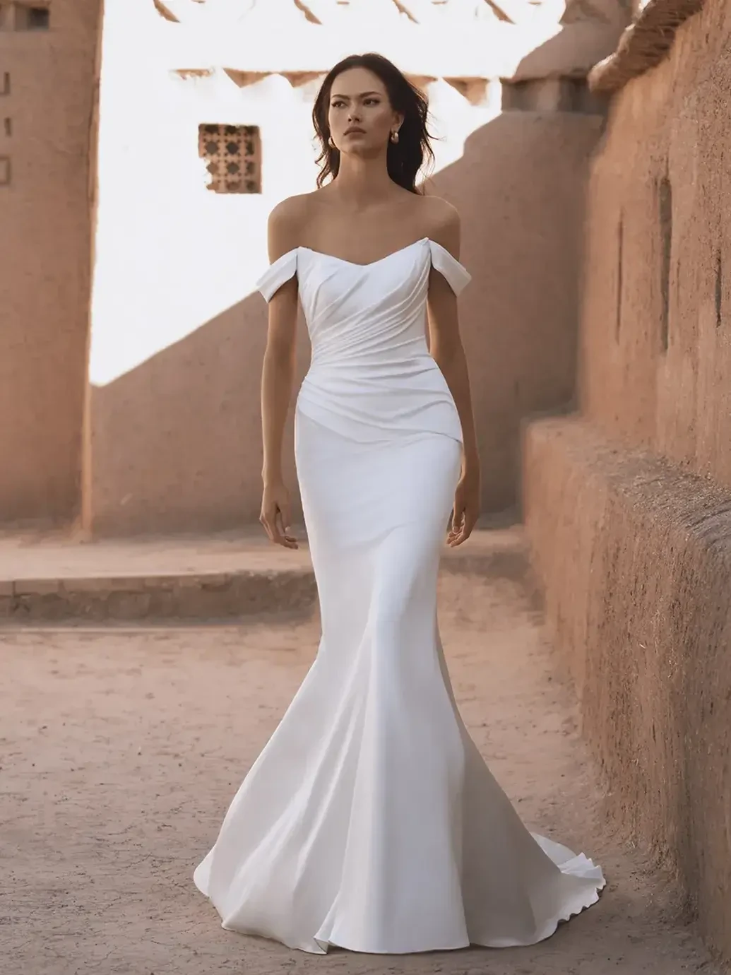 A woman in an elegant, off-shoulder white gown walks through a sunlit, narrow alley with textured clay walls, creating a serene and confident mood.