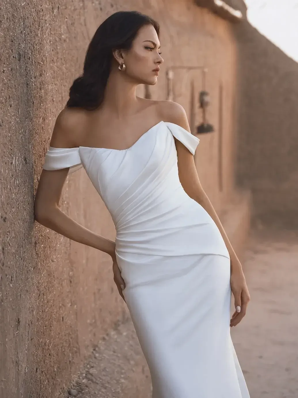 A woman in an elegant, off-shoulder white gown walks through a sunlit, narrow alley with textured clay walls, creating a serene and confident mood.