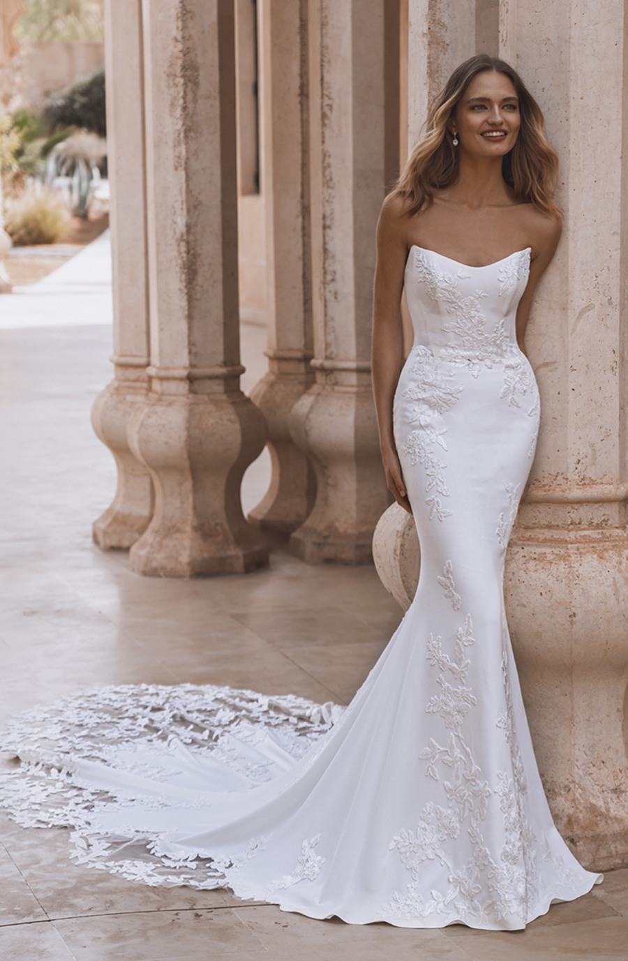 A woman in a strapless, white lace bridal gown with a long train stands elegantly by stone columns, smiling. The setting is warm and inviting.
